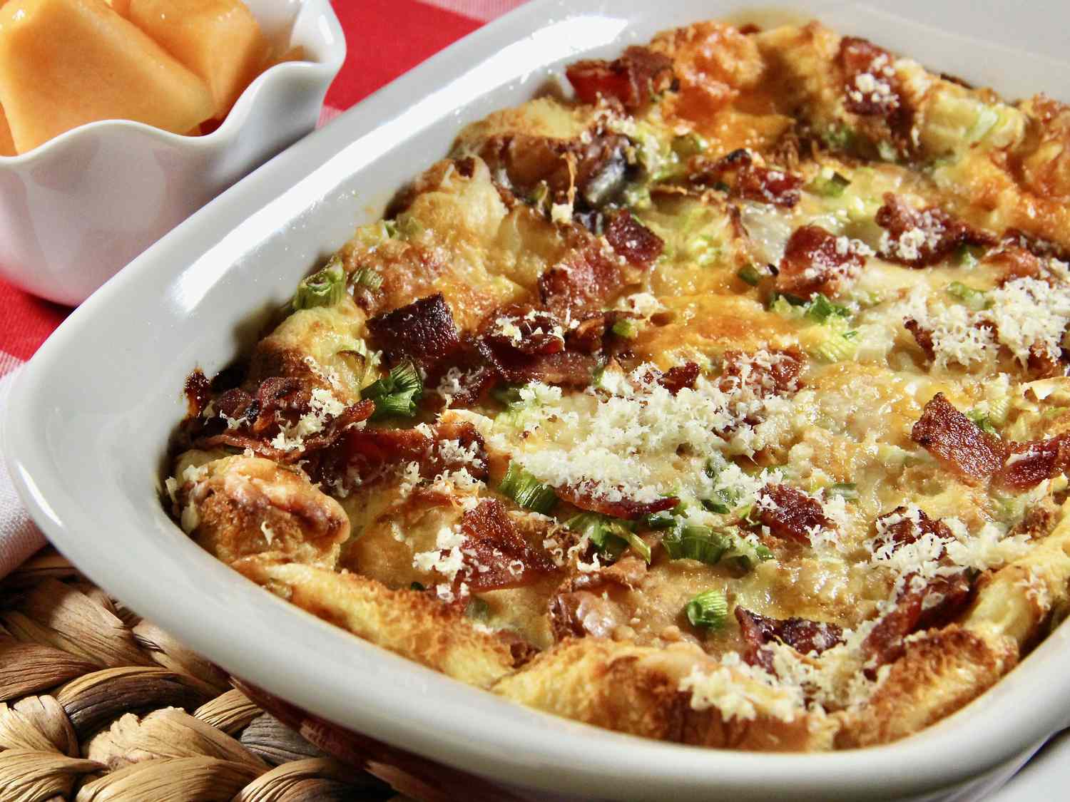 closeup of a ceramic baking dish of a breakfast casserole made with Hawaiian rolls, bacon, and cheese with a dish of cantaloupe pieces in the background